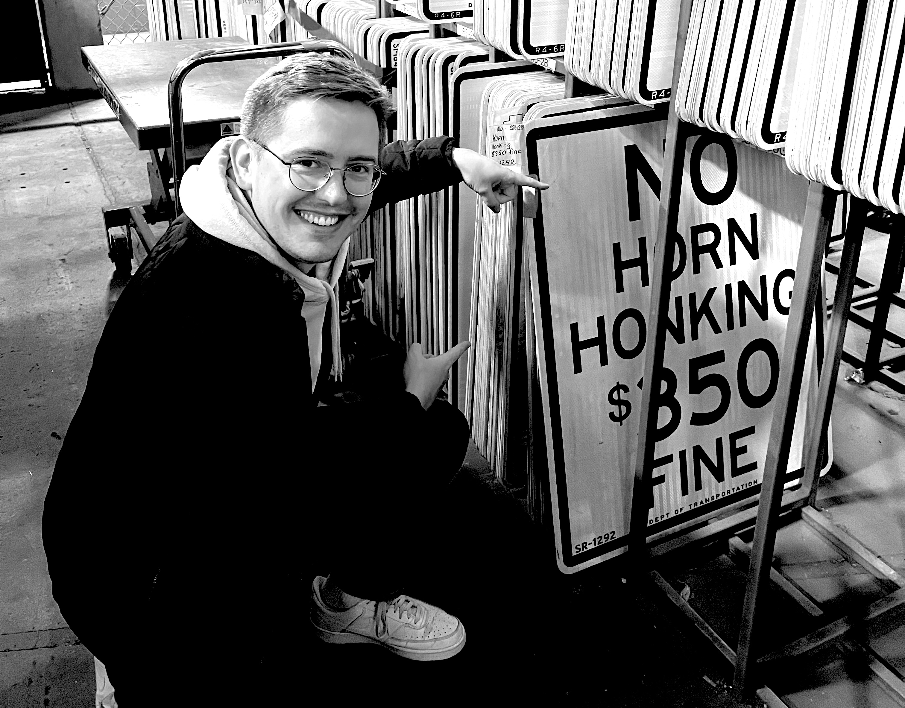 A black and white photo of Adam, a blond white man with glasses, crouched next to a rack of road signs, pointing at one that reads No Horn Honking - $350 fine