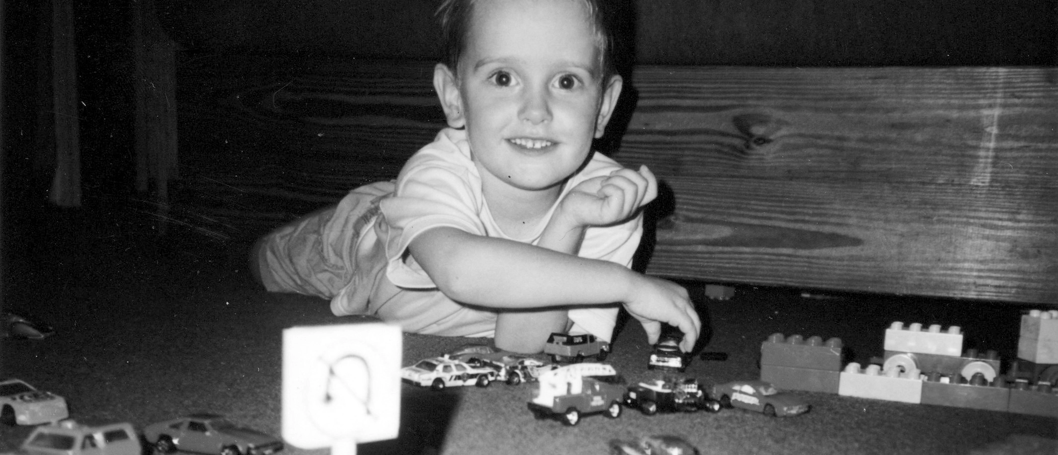 A black and white childhood photo of Adam laying on the floor playing with miniature cars